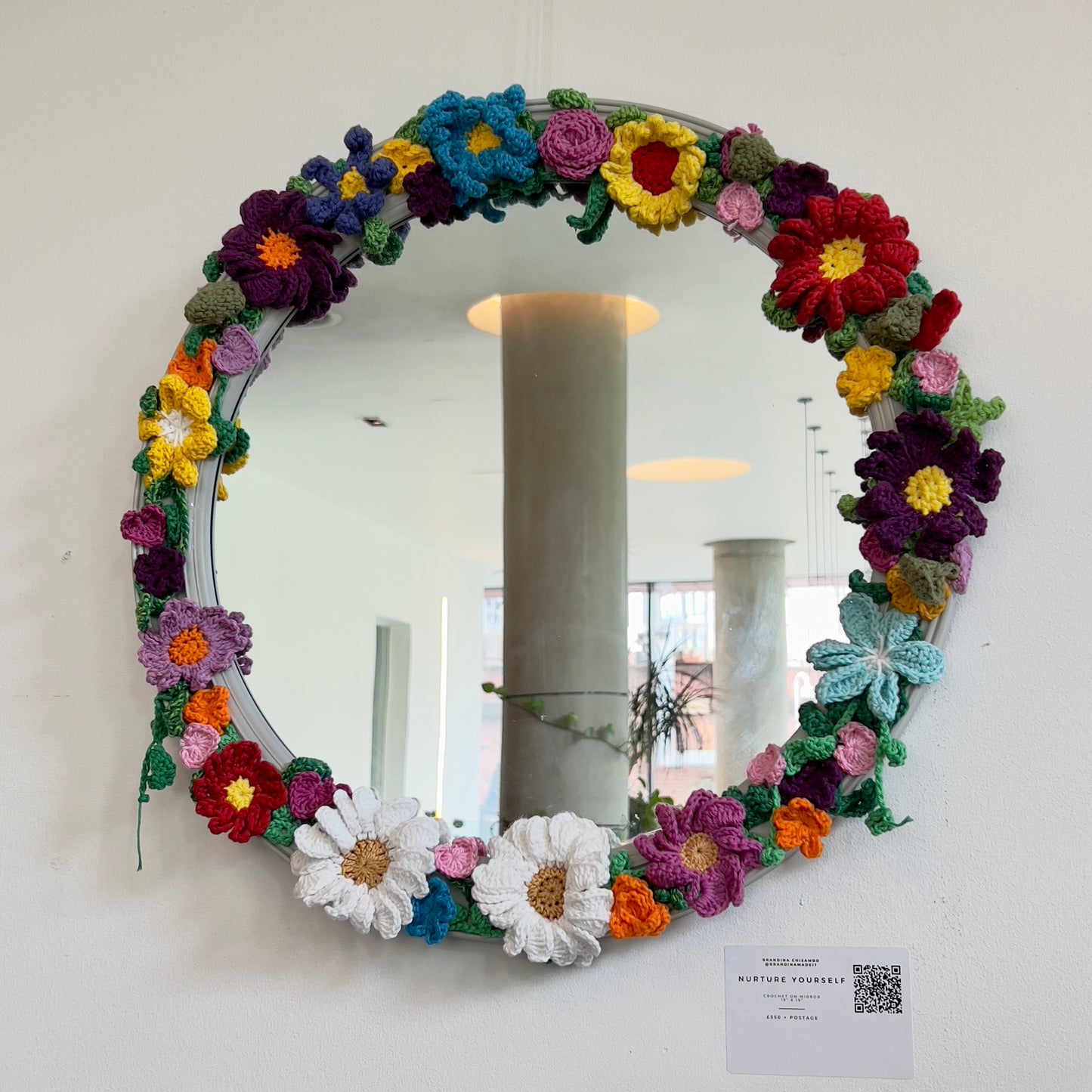 Circular wall mirror decorated with a lush, handmade wreath of colorful 3D crochet flowers including daisies, roses, and pansies, available at HEADS Art Gallery.
