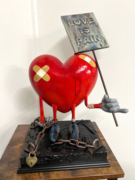 Statue of a red heart with bandages, chains, a sign reading 'Love is Pain' and chained to a base available at HEADS Art Gallery.