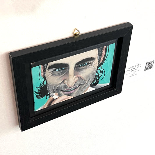 Portrait of Joaquin Phoenix as the Joker with a slight smile, holding a cigarette against a bright teal background, available at HEADS Art Gallery.