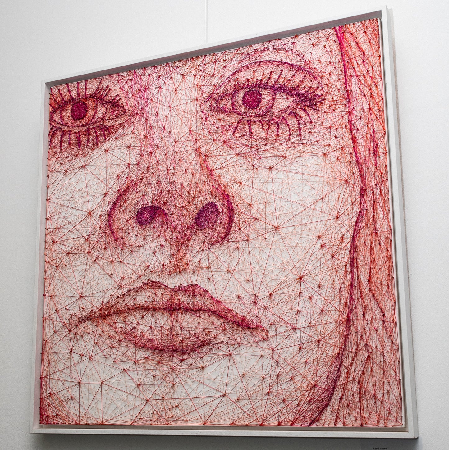 Intricate piece of string art using pink and red threads to create a detailed close-up portrait of a woman's face, available at HEADS Art Gallery.